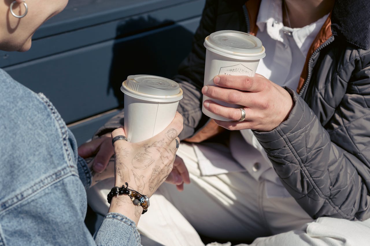 Two friends enjoying a casual coffee break outdoors with warm drinks in disposable cups.