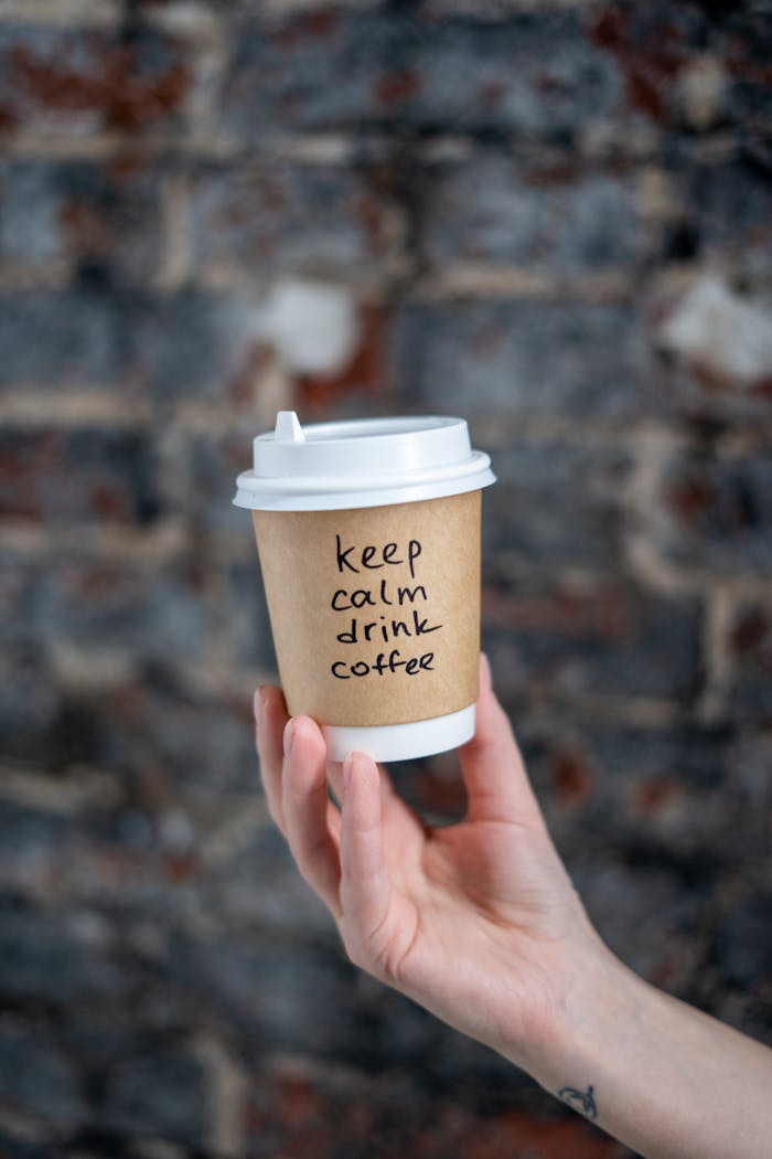 A hand holding a takeaway coffee cup with keep calm drink coffee written on it in front of a rustic brick wall.