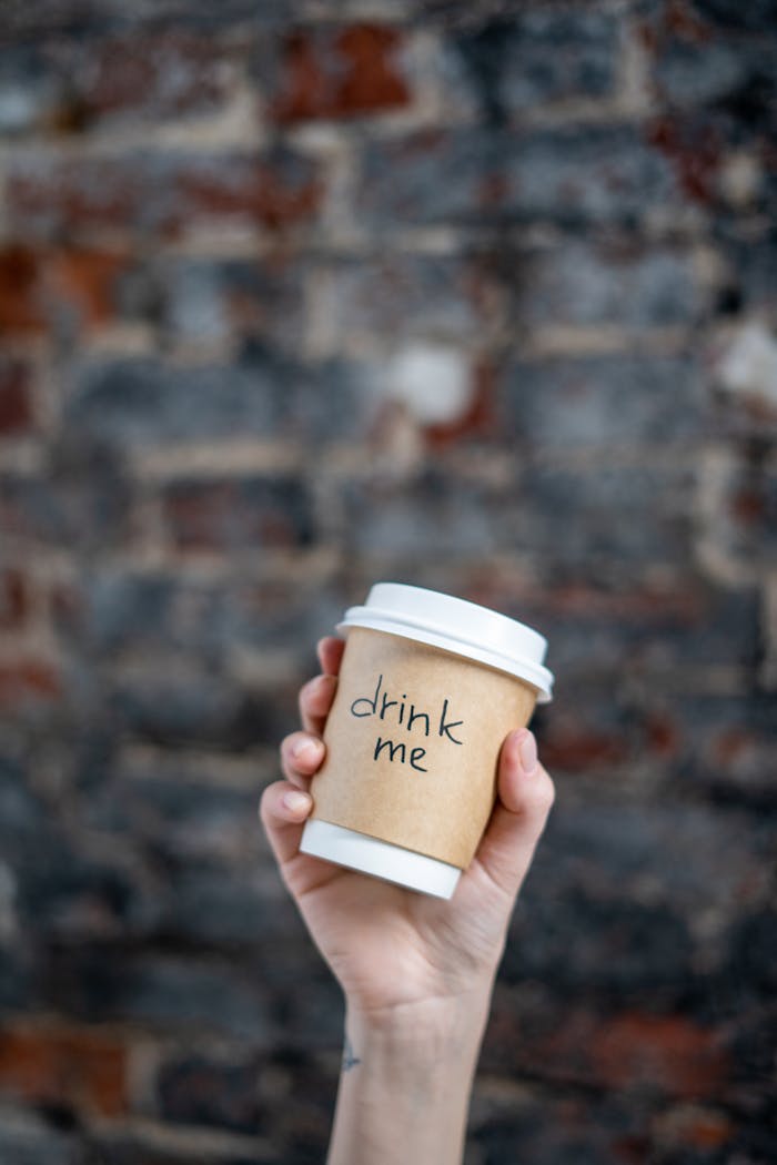 A hand holds a disposable coffee cup with drink me text against a textured brick wall.